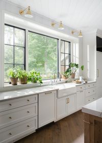 North Penn | Hoskins Interior Design, Farmhouse Sink, Painted Cabinetry and Wood Island, Quartzite Countertops, Brass Hardware, Brass Lighting, White Kitchen Cabinets, Wood Floors, Large Kitchen Window, Kitchen Plant Shelf, Black Windows