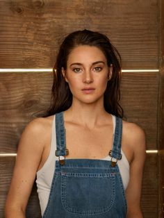 a woman in overalls is standing against a wooden wall with her hands on her hips