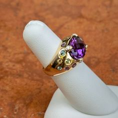 Estate/ vintage 14KT yellow gold checkerboard, oval, genuine, purple amethyst, gypsy-set multi gemstones, and diamond ring. Wide, cigar band style with vibrant colors. Size 6 Can be resized by us for a fee, or your local jeweler Ring weight: 4.90 DWT (4) Diamonds on prongs 13.8mm x 10mm center, oval, checkerboard faceted amethyst stone set in yellow gold. Gypsy-set, multi gemstones including citrine, tourmalines, etc. Stamped 14K and Hallmarked B & M Collectible Oval Rings With Gemstone Accents, Luxury Purple Multi-stone Sapphire Ring, Heirloom Purple Rings With Gemstone Accents, Oval Multi-stone Gemstones For Collectors, Multicolor Oval Amethyst Ring With Gemstone Accents, Multicolor Oval Amethyst Ring With Accent Stones, Multicolor Amethyst Ring Fine Jewelry, Heirloom Oval Sapphire Ring With Gemstone Accents, Multicolor Amethyst Rings With Accent Stones