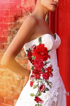 a woman wearing a white dress with red flowers on it standing in front of a brick wall