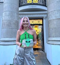 a woman with pink hair holding a cake in front of an entrance to a building