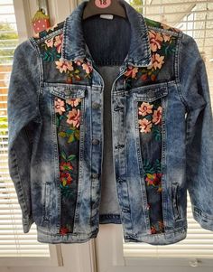 a jean jacket with embroidered flowers on the front and back, hanging from a window sill