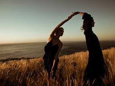 two women standing in tall grass near the ocean with their arms stretched out to each other