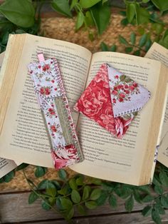 two bookmarks made out of fabric sitting on top of an open book next to green leaves