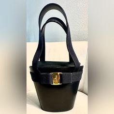 This Is A Black Calfskin Leather Handbag Made In Italy. It Features An Applied Grosgrain Ribbon And A Vara Bow Centered With A Gold Tone Ferragamo Signature Metal Buckle. The Bag Has A Magnetic Snap Closure And Opens To A Fabric-Lined Interior With A Single Zip Pocket. The Leather Top Handles Are Wrapped With Grosgrain Ribbon. The Dimensions Of The Bag Are 7 X 5 X 3 Inches, And The Drop Of The Handles Is 5 Inches. Condition: Very Good Condition. Has A Some White Marks And Minor Scratches On The Salvatore Ferragamo Bags, Small Buckets, How To Make Handbags, White Mark, Metal Buckles, Grosgrain Ribbon, Leather Handbag, Leather Top, Salvatore Ferragamo