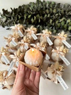 a hand holding a small candle in a box with tags around it and flowers behind it