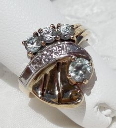 "This great, vintage two-tone ring shows detail and flair typical of the 1940s. The asymmetrical design is composed yellow gold, white gold, and four light blue topaz gemstones. The white gold \"ribbon\" is chased with tiny orange blossoms and decorated with milgrain. It separates the three smaller stones from the one larger stone. The fanned yellow gold section, which shares the side with the larger stone, has two slits and a little curl in the corner. The asymmetry even extends to the shank, w Vintage White Gold Multi-stone Ring, Vintage Multi-stone Diamond Ring Collectible, Vintage Multi-stone Diamond Ring For Collectors, Vintage White Gold Multi-stone Diamond Ring, Vintage Multi-stone Diamond Ring In White Gold, Vintage White Gold Topaz Ring With Round Cut, Vintage Topaz Ring With Round Cut For Anniversary, Vintage White Gold Topaz Ring With Center Stone, Vintage Round Cut Topaz Ring For Anniversary