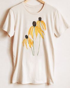 a white t - shirt with yellow flowers painted on the front and back, hanging from a wooden hanger