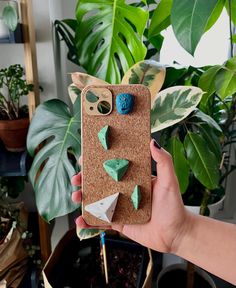 a person holding up a phone case with green and blue decorations on the back, in front of a potted plant