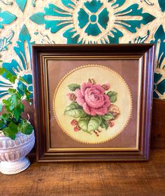 a cross stitch rose in a frame next to a potted plant on a table