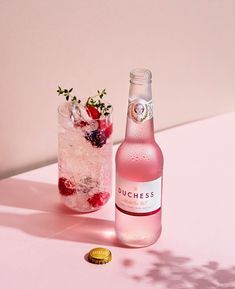 a bottle of punch next to a glass filled with ice and raspberries on a pink table