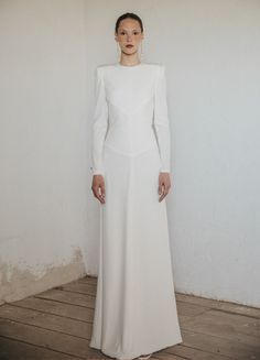 a woman standing in front of a white wall wearing a long white dress and earrings