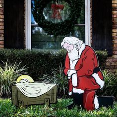 a santa clause sitting in the grass next to a toy bed and chest with a christmas wreath on it