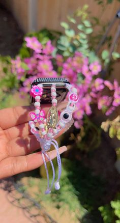 a hand holding a cell phone in front of some pink flowers and purple flowers on the ground