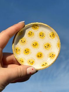 a hand holding a yellow and white paper plate with smiley faces on it, against a blue sky