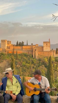 three men are sitting on a wall playing guitar and singing in front of a castle