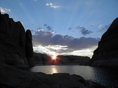 the sun is setting in between two large rocks on the water's edge, with mountains and cliffs behind it