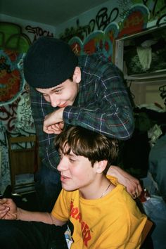 two young men sitting next to each other in front of a wall covered with graffiti