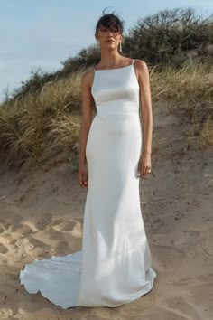 a woman standing in the sand wearing a white wedding dress with one shoulder cut out