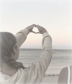 a woman making a heart shape with her hands on the beach near the ocean at sunset