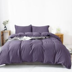 a bed with purple sheets and pillows in a white room, next to a nightstand