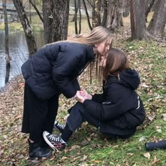 two women kneeling down to kiss each other in front of some trees and water with leaves on the ground