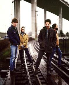 four men standing on train tracks in front of an overpass with one man leaning against the rail