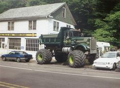 a large green truck parked in front of a white building next to a forest filled with trees