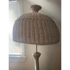 a wicker table lamp with a white shade on the top and bottom, in front of a window