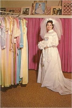 a woman in a wedding dress standing next to dresses hanging on a rack and smiling at the camera