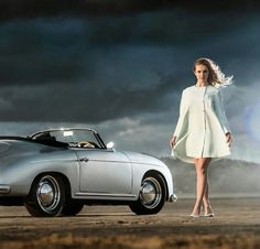 a woman standing next to a silver car in the middle of an empty field with dark clouds