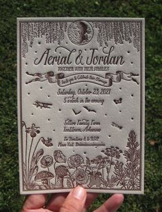 a hand holding up a wedding card in front of green grass with trees and animals on it