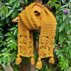 a yellow crocheted scarf with tassels hanging on a wooden stand in front of some bushes