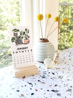 a desk with a calendar and flowers in a vase next to a window sill