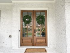 two wreaths on the front door of a white brick house with welcome mat in front