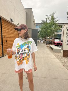 a woman holding a drink and wearing mickey mouse t - shirt while standing in front of a building