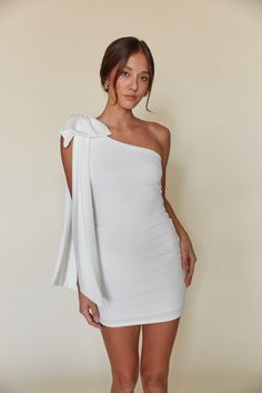 Turn heads in this sweet and special mini dress. The Mischa One Shoulder Bow Bodycon Mini Dress features a bodycon silhouette, one shoulder neckline with a sweeping bow detail. Available in white, black, and red. Pair with a sleek heel to complete the look! Details 96% Polyester, 4% Spandex Lining: 100% Polyester Fully lined Hand wash cold / Line dry White Dress With Red Accessories, One Shoulder White Dress Short, Graduation Dresses Winter, Hoco Dresses With Bow, Short Grad Dress, White Halter Dress Short, White Hoco Dress, White Dress With Bow, White Semi Formal Dress