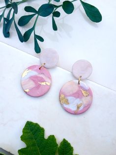 the pink and gold earrings are next to a green plant on a white surface with leaves