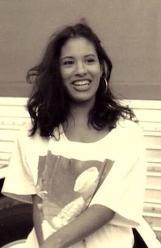 a black and white photo of a smiling woman in a t - shirt sitting on a bench