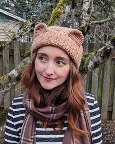 a woman wearing a knitted hat and scarf in front of a fence with trees