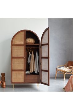 an open wooden cabinet with clothes hanging on hooks and two wicker chairs in the background