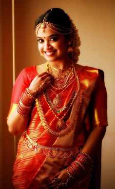 a woman in an orange and red sari with gold jewelry on her neck, smiling at the camera