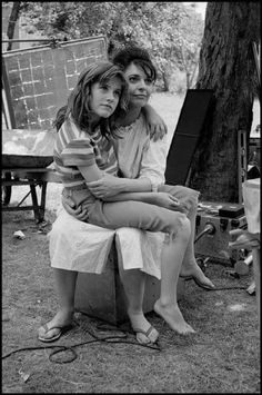 two young women sitting next to each other in the grass near a tree and an easel