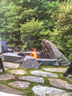 an outdoor fire pit surrounded by rocks and grass with trees in the backround