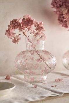 a glass vase with pink flowers in it on a white table cloth next to two plates