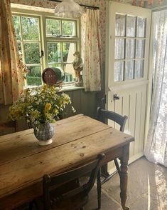 a wooden table sitting in front of a window next to a chair and vase with flowers on it