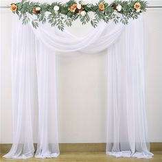 a white drape with flowers and greenery on the top is hanging in front of a wall