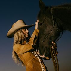 a woman wearing a cowboy hat is petting a horse's nose while standing in front of a dark blue sky