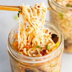 two jars filled with noodles and vegetables being held by chopsticks