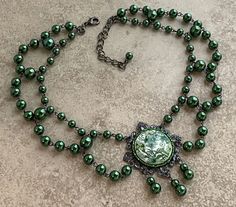 The centerpiece for this vintage-style necklace is a round floral print cabochon in shades of green on white, mounted on a square gunmetal filigree.  Wire-linked forest green glass pearls in 3 sizes  are festooned together to about 14"; it finishes with a lobster clasp and length of chain for adjustability, so may be worn from about 15" to 18"  (38-45cm).  I can add or subtract length if you need it--just let me know when you order! In the pictures the pearls might appear a bit olivine, but they are a really satisfying forest green. Festoon Necklace, Vintage Style Necklace, Edwardian Style, Edwardian Fashion, Choker Necklaces, Style Necklace, Vintage Stil, Green Glass, Shades Of Green